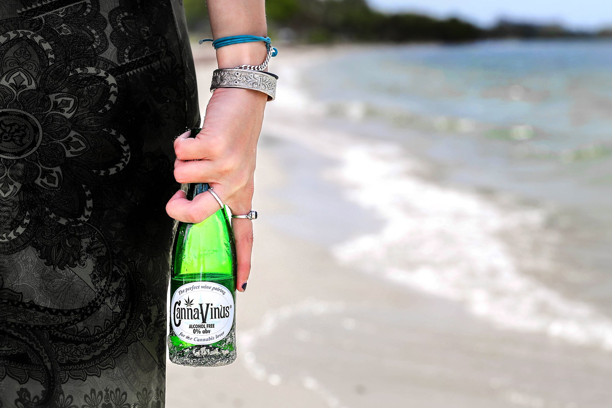 an image of a woman holding a CannaVinus bottle on a beach with water in the background